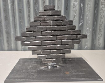 May include: A diamond-shaped metal sculpture composed of rectangular blocks, set on a rectangular base. The sculpture is made of dark grey metal. The background features a corrugated metal sheet.