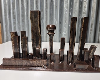 May include: A dark brown metal sculpture of a cityscape. It features rectangular and cylindrical shapes of varying heights, arranged on a rectangular base. The sculpture has a rustic, industrial aesthetic, with a focus on geometric forms.