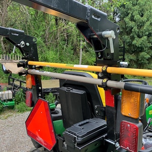 May include: A black metal tractor attachment rack mounted on the rear of a green tractor, featuring two horizontal arms holding a yellow rake handle and a grey tool handle, with red and orange safety reflectors and a triangular slow-moving vehicle sign.