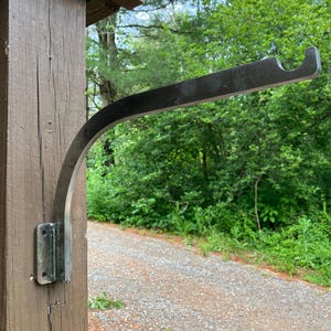May include: A metal bracket, likely for hanging a plant or other item, is attached to a wooden post. The bracket has a curved arm and a hook-like end. The post is brown, and the background shows a gravel path and green foliage.