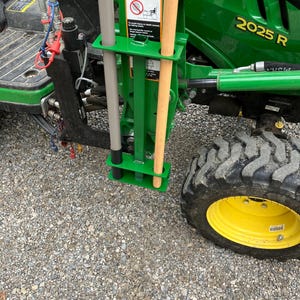 May include: A green metal tool holder attached to a John Deere 2025 R tractor. The holder contains a gray metal bar and a wooden-handled tool. A warning sign is visible on the holder.