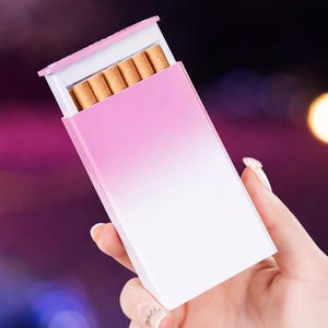 Może przedstawiać: Różowy i biały gradientowy futerał na papierosy, częściowo otwarty, aby odsłonić papierosy. Etui jest trzymane w dłoni na rozmytym tle ze światłami bokeh.