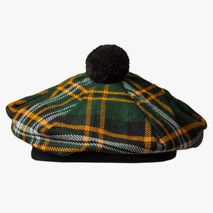 May include: A green, yellow, and white tartan hat with a black pom-pom. The hat has a black brim and a flat shape. This hat is a traditional style, often worn in Scotland.
