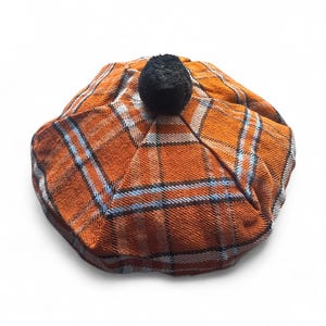 May include: An orange plaid beret with a black pom-pom. The beret has a classic plaid pattern in orange, white, and blue. This hat is a stylish accessory.
