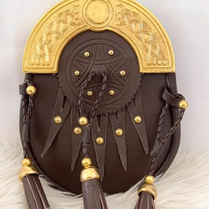 Puede incluir: Una sporran de cuero marrón con una parte superior y detalles de metal dorado. La sporran presenta un diseño de nudo celta decorativo, tachuelas doradas y tres borlas con tapas doradas. Un accesorio escocés tradicional.