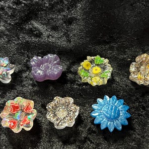 May include: Seven colorful glass flower buttons with various designs and textures. The buttons are iridescent, glitter, and opaque.