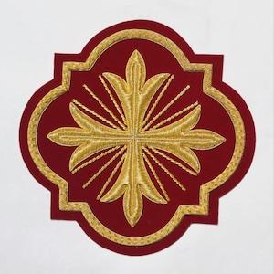 Puede incluir: Un parche de color rojo oscuro con forma de cuatrifolio que presenta un intrincado bordado dorado. El centro muestra una cruz estilizada con terminales de flor de lis, rodeada de rayos solares, todo dentro de un borde dorado decorativo.