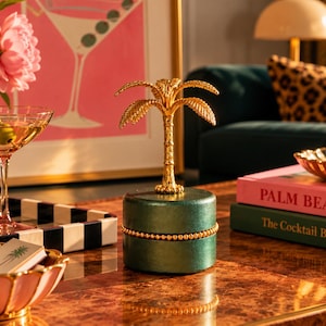 May include: A cylindrical dark green jewellery box with a gold beaded trim around the centre and a gold metal palm tree figurine attached to the lid. The box has a smooth, matte finish and stands upright as a decorative storage container for small items.