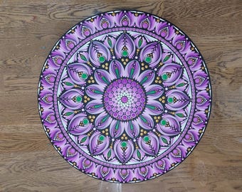 May include: A round, hand-painted mandala in shades of purple, green, and gold. The intricate design features a central floral pattern surrounded by layers of detailed, symmetrical elements. The artwork is set against a black background.