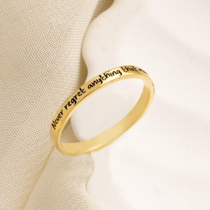 以下が含まれることがあります： 「Never regret anything that」の黒い文字が刻まれたゴールドカラーのリング。白い波型の表面とベージュの布の背景にセットされています。