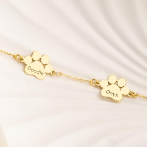 Puede incluir: Una pulsera dorada con dos colgantes con forma de huella de pata. Cada colgante tiene un nombre grabado: "Doodle" y "Onyx". La pulsera se muestra sobre un plato blanco con forma de concha y tela crema.