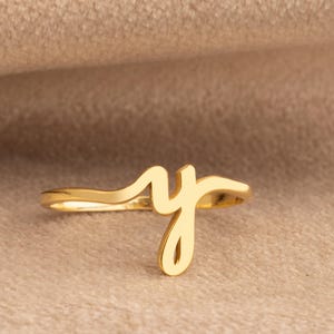 May include: A gold-colored ring featuring the cursive letter "y". The ring is set against a neutral, tan-colored background. The ring is a delicate piece of jewelry.