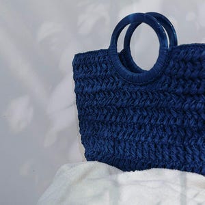 May include: A navy blue woven tote bag with two circular handles. The bag has a textured, basket-weave appearance. The handles are a darker shade of blue. The bag is sitting on a white, fluffy surface.