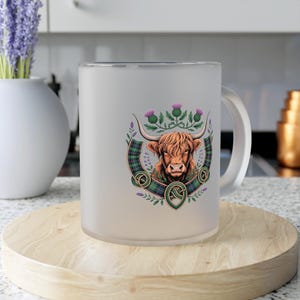 May include: Frosted glass mug with a Highland cow design. The image shows a brown cow with long horns, set against a green and black tartan pattern, purple thistles, and Celtic knot details. The mug has a curved handle and is on a wooden coaster.