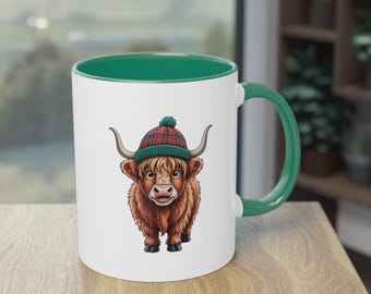 May include: White ceramic mug with a green interior and handle. The mug features a cartoon illustration of a Highland cow wearing a plaid hat. The cow is brown with long fur and curved horns. The mug is on a wooden surface.