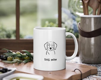 Puede incluir: Taza de cerámica blanca con un dibujo lineal negro de la cara de un perro y el texto "DOG MOM". La taza está sobre una tabla de madera. El fondo incluye una ventana y un recipiente con cucharas de madera.