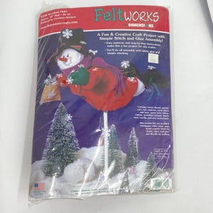 以下が含まれることがあります： FeltWorks by Dimensionsのクラフトキット「Snowflake Flight」。パッケージには、黒い帽子、赤いコート、紫色のマフラーを身につけた雪だるまが、緑色のボトルブラシツリーの間で白い雪の土台の上に立っている様子が描かれています。