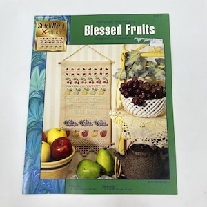 May include: A cross-stitch pattern book titled "Blessed Fruits" with a design of fruits and the text "A Good Tree Cannot Bear Bad Fruit." The cover features a finished cross-stitch piece, a bowl of apples, and a basket of grapes.