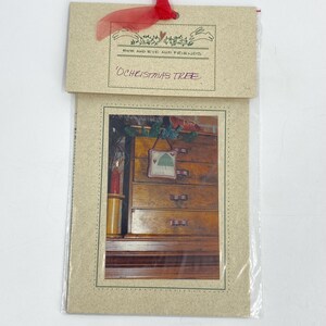 May include: A craft pattern packet titled Ewe and Eye and Friends O Christmas Tree featuring a printed image of a small square hanging ornament with a green tree design, displayed on a wooden chest of drawers with a red candle nearby.