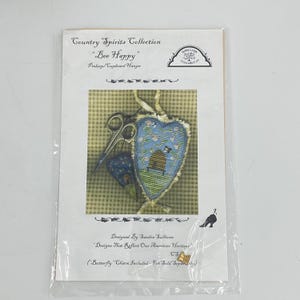 May include: A Country Spirits Collection Bee Happy pinkeep and cupboard hanger pattern packet. The cover features a heart-shaped blue cross-stitch design with a beehive, scissors, and a small gold butterfly charm included in the packaging.