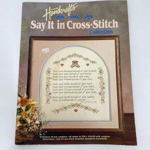 May include: A vintage "Say It in Cross-Stitch" collection book. The cover features a framed cross-stitch sampler with a poem. The book's title is in a decorative font, and the cover has a floral arrangement in the lower left corner.