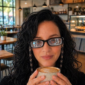 May include: A woman with dark curly hair wearing black-framed glasses with a gold chain. She holds a small, light-coloured ceramic cup of coffee with a foamy top. The background shows a cafe setting with tables and hanging lights.