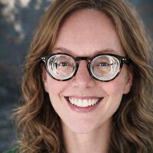 May include: A person smiling, wearing round tortoiseshell glasses with thick frames and clear lenses. The person has light brown wavy hair and is wearing a green shirt. The background is blurred with shades of blue and grey.