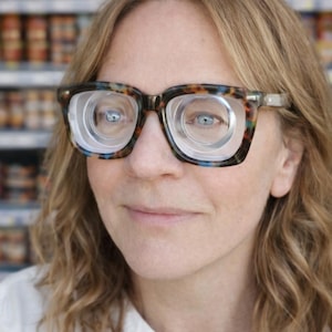 May include: A woman wearing glasses with a tortoiseshell pattern frame. The lenses feature a unique concentric circle design. The background is blurred, suggesting a grocery store setting. The woman has fair skin and light brown wavy hair.