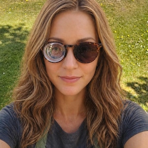 May include: Sunglasses with a tortoiseshell frame, featuring a dark tinted lens and a clear, magnified lens. The person is wearing a grey t-shirt and a green apron. The background is a grassy area with sunlight.