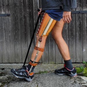 May include: A leg brace featuring a tan leather thigh cuff with white lacing, connected to a metal frame with brown leather straps around the knee and calf. The brace is worn on one leg, paired with a black walking cane and athletic sneakers.