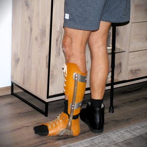 Puede incluir: Una &oacute;rtesis de pierna de color marr&oacute;n y plateado con un zapato a juego. La &oacute;rtesis se sujeta a la pierna con soportes met&aacute;licos y cordones. El zapato es negro. La persona lleva pantalones cortos grises y calcetines negros.