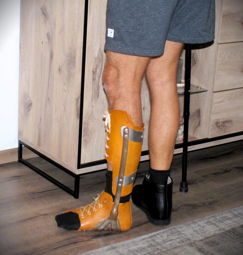 Puede incluir: Una &oacute;rtesis de pierna de color marr&oacute;n y plateado con un zapato a juego. La &oacute;rtesis se sujeta a la pierna con soportes met&aacute;licos y cordones. El zapato es negro. La persona lleva pantalones cortos grises y calcetines negros.