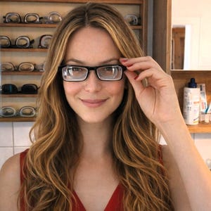 May include: A woman wearing black rectangular eyeglasses. She is smiling and holding the frame of the glasses with her right hand. A wooden shelf with various pairs of spectacles is visible in the background. The woman is wearing a red top.