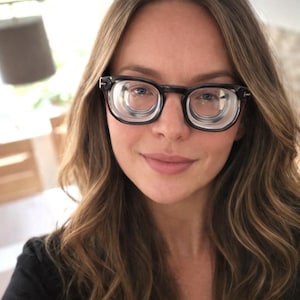 May include: Eyeglasses with a black frame and a distinctive circular lens design. The glasses have a classic rectangular shape with a thick frame. The lenses appear to have a double-lens construction. The person in the image is smiling.
