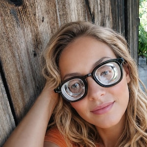 May include: Black-framed eyeglasses featuring thick, rounded rectangular rims and prominent, highly convex magnifying lenses. The glasses have a sturdy bridge and temples, designed with a classic aesthetic that emphasizes the depth of the lenses.
