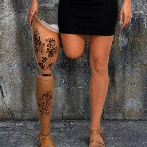 Puede incluir: Una pierna protésica con diseños florales se muestra junto a una persona que lleva una falda negra y sandalias. La pierna protésica es de color canela con tatuajes florales negros. La persona está de pie sobre una superficie de hormigón.