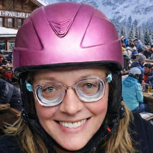May include: A magenta ski helmet with a black chin strap and clear eyeglasses. The person is smiling, with a snowy mountain backdrop and a wooden building in the background.