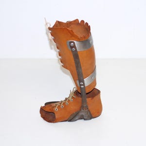 Puede incluir: Una antigua ortesis de pierna de cuero marr&oacute;n con soportes y detalles met&aacute;licos. La ortesis presenta una secci&oacute;n superior con cordones y una parte inferior en forma de zapato. Las bandas y soportes met&aacute;licos est&aacute;n remachados al cuero.