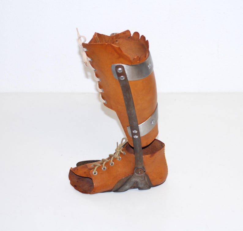 Puede incluir: Una antigua ortesis de pierna de cuero marr&oacute;n con soportes y detalles met&aacute;licos. La ortesis presenta una secci&oacute;n superior con cordones y una parte inferior en forma de zapato. Las bandas y soportes met&aacute;licos est&aacute;n remachados al cuero.