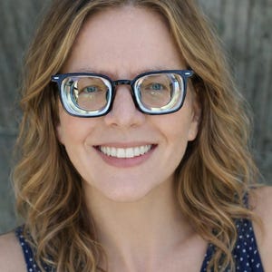 May include: A person smiling, wearing black-framed spectacles. They have fair skin and wavy blonde hair, and are wearing a blue and white polka dot top. The background is a blurred grey.