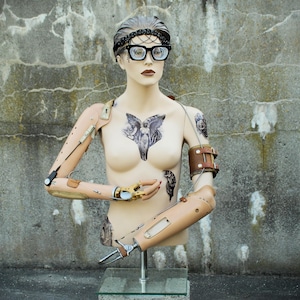 May include: A mannequin torso with mechanical arm replacements and tattoos. The mannequin wears black-framed glasses, a jeweled headband, and a brown leather arm cuff. The torso is displayed on a glass stand against a textured concrete wall.