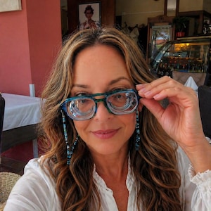 May include: A woman wearing blue and green eyeglasses with a beaded eyeglass chain. The eyeglasses have a clear lens and a thick frame. The woman is wearing a white blouse. The background is a restaurant.