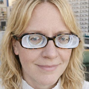 May include: A woman wearing glasses with thick, tortoiseshell frames. The lenses have a unique, concentric design, creating a magnified effect. She is wearing a white collared shirt, and her blonde hair is shoulder-length.