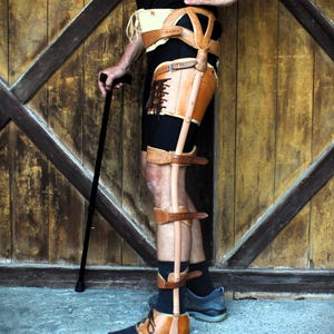 Puede incluir: Una órtesis de pierna de cuero marrón con correas y hebillas, que se extiende desde la cadera hasta el tobillo. La órtesis se lleva sobre pantalones cortos y calcetines negros, con un zapato marrón y negro. Se sostiene un bastón negro.