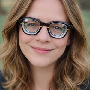 May include: A woman wearing black-framed spectacles with a yellow accent on the temples. The spectacles have a unique, modern design. The woman has light brown wavy hair and is smiling. The background is blurred, suggesting an outdoor setting.