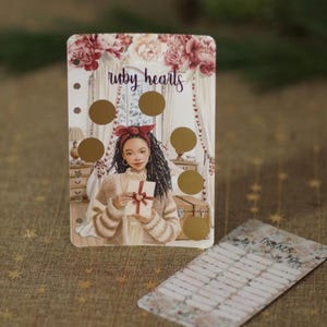 Könnte beinhalten: Eine Planungskarte mit dem Text "ruby hearts" und einer Illustration einer Frau, die ein Geschenk hält. Die Karte hat goldene Rubbelkreise und eine kleinere Tracker-Karte mit floralen Akzenten. Die Farbpalette umfasst Beige, Rosa und Gold.