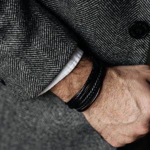 Może przedstawiać: Zbliżenie czarnej skórzanej bransoletki z wieloma pasmami, noszonej na nadgarstku. Bransoletka ma plecione i gładkie tekstury. Tło przedstawia szary płaszcz w jodełkę i biały mankiet koszuli.