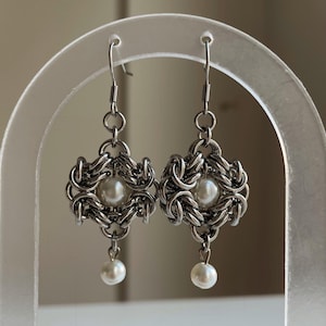 Może przedstawiać: Para srebrnych kolczyków. Każdy kolczyk ma pleciony metalowy wzór z centralną perłą i wiszącą perłą. Kolczyki są prezentowane na przezroczystym, łukowatym stojaku na neutralnym tle. Design jest skomplikowany i elegancki, idealny na specjalne okazje.