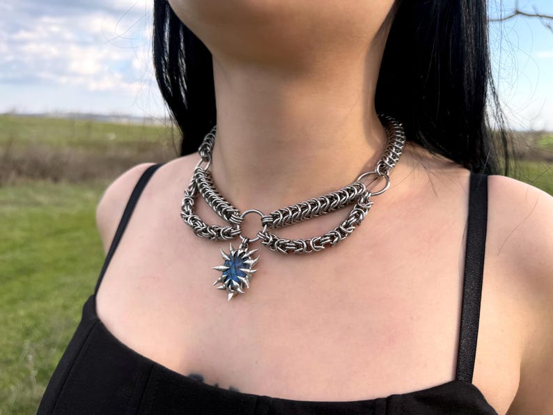 May include: A silver-toned chainmail necklace with a blue gemstone pendant. The necklace features multiple layers of chain and a central pendant shaped like a starburst. The gemstone has a dark blue color and is surrounded by pointed metal accents.