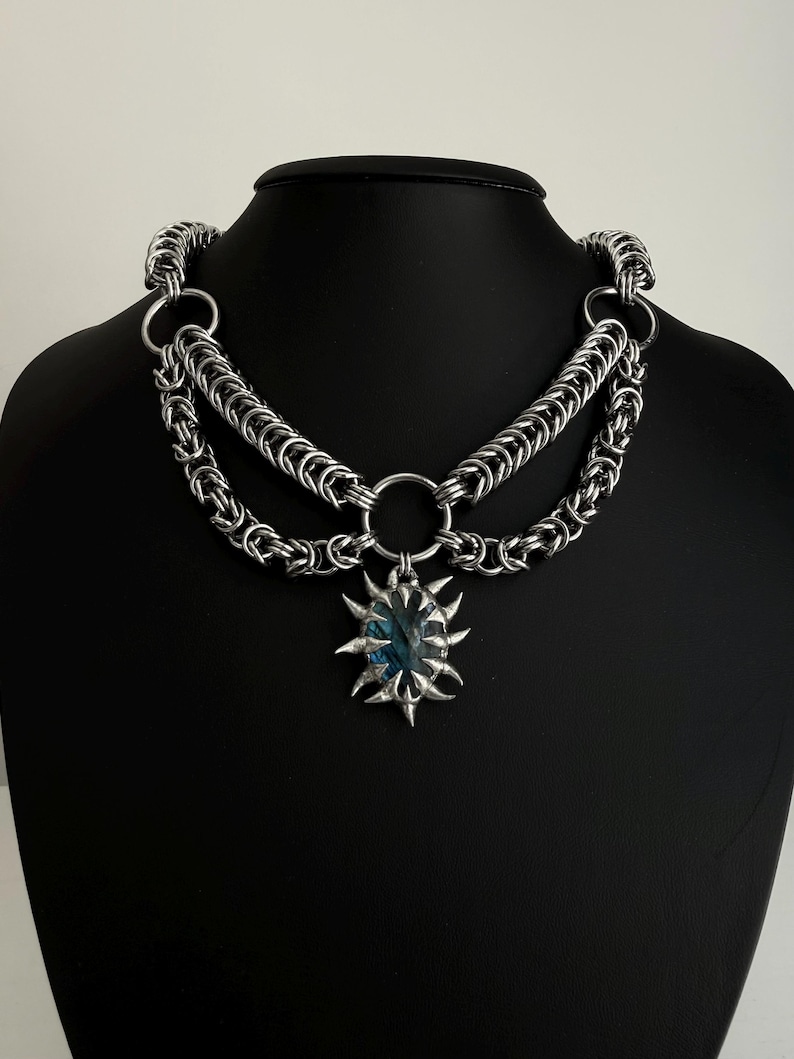 May include: A silver-toned chainmail necklace with two layers of chain and a central pendant. The pendant is a sunburst design with a blue-green stone at its center. The necklace is displayed on a black mannequin.